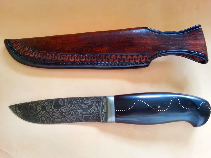 Wave-pattern Damascus knife with a dark horn handle, silver-wire inlay, and a shaped brown leather sheath