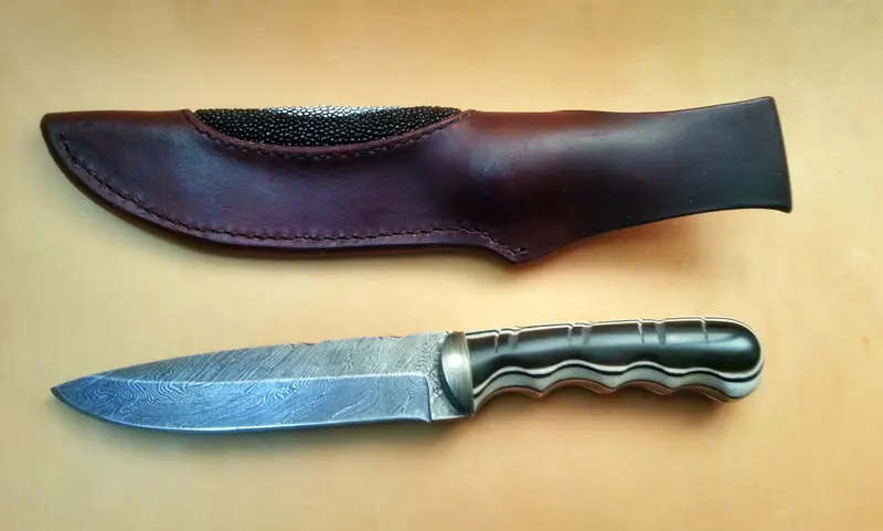 Patterned-blade knife with a sculpted dark handle and a burgundy leather sheath