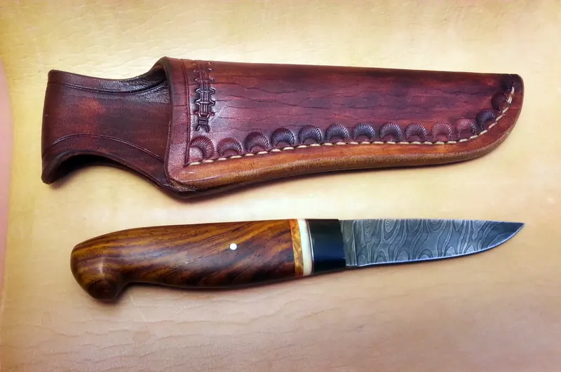 Knife with a patterned blade and curved wooden handle alongside its shaped brown leather sheath